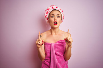 Sticker - Young beautiful woman wearing towel and bath hat after shower over pink isolated background amazed and surprised looking up and pointing with fingers and raised arms.