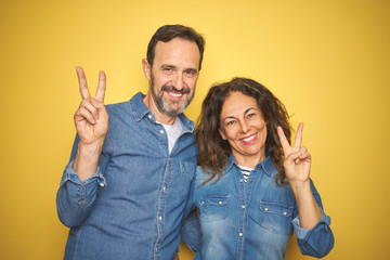 Beautiful middle age couple together standing over isolated yellow background smiling looking to the camera showing fingers doing victory sign. Number two.