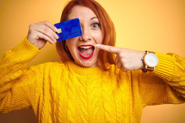 Poster - Young beautiful redhead woman holding credit card over yellow isolated background very happy pointing with hand and finger