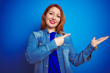 Sticker - Young beautiful redhead woman wearing denim shirt standing over blue isolated background amazed and smiling to the camera while presenting with hand and pointing with finger.