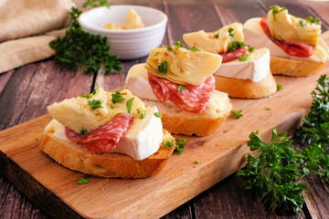 Crostini appetizers with brie cheese, salami and artichokes. Close up on a serving board against a wood background. Party food concept.