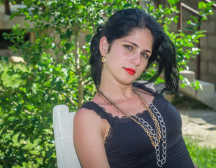 Cute Cuban girl in the yard of her house, sitting looks at camera. White skin, green eyes and red lips