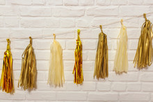 Gold Paper Garlands On A White Brick Wall.  Tissue Paper Tassels Garland Bunting Happy Birthday Wedding Party Decor. Matte And Glossy Gold Garland For Party Decoration