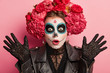 Close up shot of shocked woman wears frightening makeup, keeps palms raised, celebrates Halloween or Mexican Day of Death, isolated over pink background, scared by something. Art and face painting