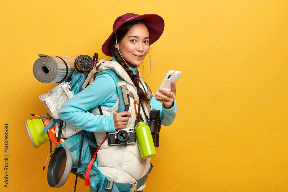 Pretty satisfied traveler uses free internet connection on smartphone for blogging during wanderlust trip, carries big heavy rucksack, has binoculars and retro camera to explore surroundings - obrazy, fototapety, plakaty 
