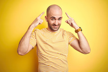 Young Bald Man With Beard Wearing Casual Striped T-shirt Over Yellow Isolated Background Smiling Pointing To Head With Both Hands Finger, Great Idea Or Thought, Good Memory