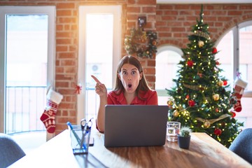 Poster - Beautiful woman sitting at the table working with laptop at home around christmas tree Surprised pointing with finger to the side, open mouth amazed expression.