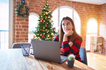 Poster - Beautiful woman sitting at the table working with laptop at home around christmas tree with hand on chin thinking about question, pensive expression. Smiling with thoughtful face. Doubt concept.