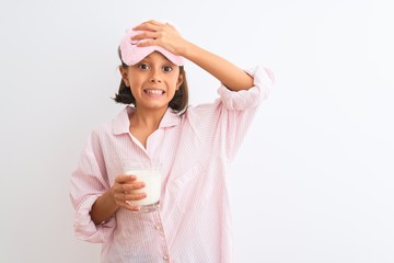 Poster - Child girl wearing sleep mask and pajama drinking glass of milk over isolated white background stressed with hand on head, shocked with shame and surprise face, angry and frustrated. Fear and upset