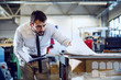 © Dusan Petkovic - Dedicated Caucasian bearded graphic engineer in shirt and tie holding notebook and looking at sheets while standing in print shop.