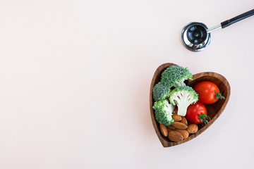 Healthy food for heart on light background. Plate with vegetables, nuts and a medical stethoscope on the table. Diet and medicine concept. Top view, copy space