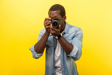 Portrait of male photographer or traveler in denim casual shirt holding professional digital dslr camera and focusing, taking photo, shooting video. indoor studio shot isolated on yellow background