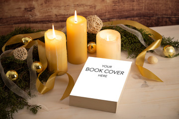 Christmas setting for a book presentation: book with blank cover settled with three lit candles , white organza and gold satin ribbons, gold baubles on light wooden table and dark wood background