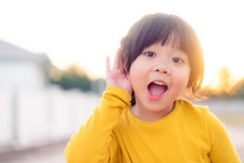 Little Asian Girl Holds Her Hand Near Her Ear And Listening Something.Exciting Face On Funny Child Girl Wear Yellow Shirt In Winter Time And Listening To Curious Good News.
