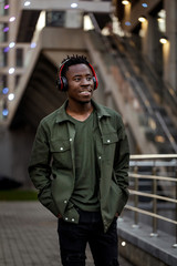 Wall Mural - smiling african-american man in stylish jacket in wireless headphones listening music on the street of the evening city