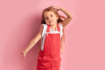 dreamy beautiful caucasian little girl in red overalls with long hair looking at camera isolated over pink background