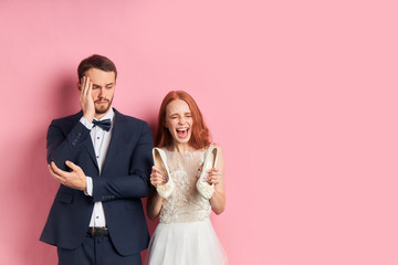 Wall Mural - Portrait of positive redhaired woman in suit seriously looking at smiling female in white wedding dress. isolated over pink background