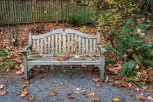 Weathered Wooden Bench Beside A Gravel Path In A Park, Fall Landscape Covered In Fallen Leaves
