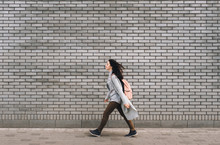 Young Beautiful Caucasian Woman In Gray Pullover With A Pink Bag Walking On The Autumn Street. Brick Wall.