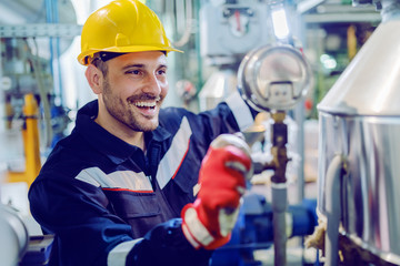 Hardworking smiling blue collar worker in protective working suit and with helmet adjusting temperature on boiler while standing in factory and holding lamp.