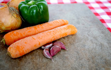  Selective Focus Of Various Vegetables Including Carrots, Brown Onions, Garlic Cloves And A Green Pepper On A Mottled Brown Board On A Red Checked Table Cloth