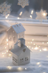 Christmas decoration, bokeh light and two little white Christmas ornaments houses on the white blanket by the white window at night. Winter wonderland decoration. Calendar, advent and Christmas card c