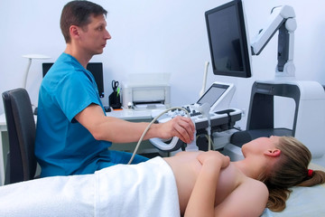 Wall Mural - Man doctor making ultrasound diagnostic of mammary glands of young woman on bust. He runs ultrasound sensor over patient's mammary glands and looks at image on screen. Diagnosis of internal organs.