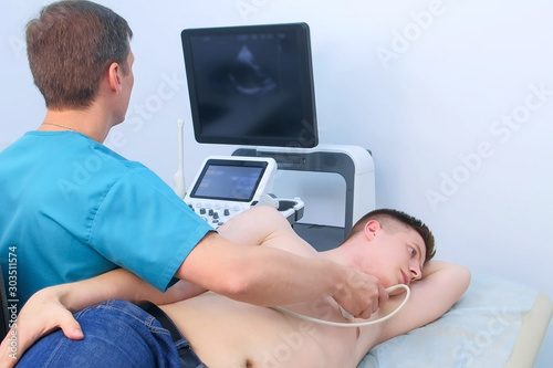 Echocardiography. Man doctor examining guy patient\'s heart by using ultrasound equipment. He runs ultrasound sensor over man\'s chest, working on scanner panel, looking at screen.