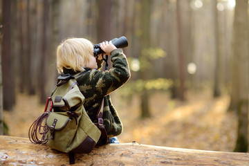 Little boy scout with binoculars during hiking in autumn forest. Child is sitting on large fallen tree and looking through a binoculars.