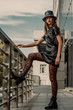 Outdoor full body street fashion portrait of young elegant confident model, woman wearing trendy bucket hat, short faux leather dress, black leopard print tights, lace up ankle military boots