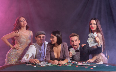 Friends playing poker at casino, at table with stacks of chips, money, cards on it. Celebrating win, smiling. Black, smoke background. Close-up.