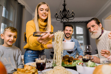 Wall Mural - Happy smiling family, grandfather, parents and children sitting at festive table and eating tasty dinner. Mother puts pasta spaghetti on plates for all family members