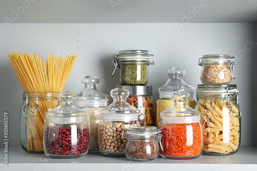 Photographie Glass jars with different types of groats and pasta on white shelf