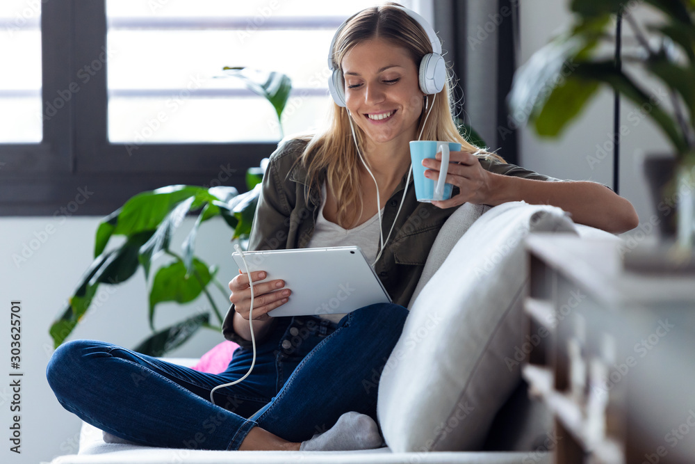 Pretty young woman listening to music with her digital tablet while drinking cup of coffee on sofa at home. - obrazy, fototapety, plakaty 