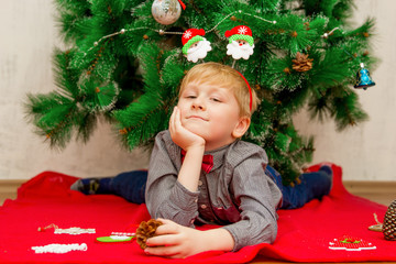 Happy smiling light haired little boy lying on a red blanket under Christmas New Year's tree with Christmas decorations around him