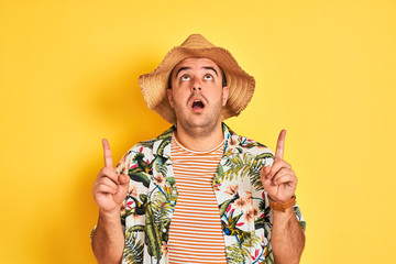 Sticker - Young man on vacation wearing summer shirt and hat over isolated yellow background amazed and surprised looking up and pointing with fingers and raised arms.