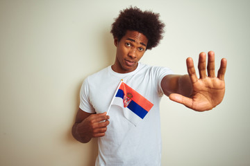 Poster - Young afro american man holding Serbia Serbian flag standing over isolated white background with open hand doing stop sign with serious and confident expression, defense gesture