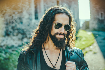 Self confident handsome man in sunglasses. Macho man walking outside alone. Stylish young fashionable man with long wavy hair wearing bikers leather jacket and aviator sunglasses.