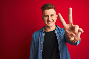 Young handsome man wearing denim shirt standing over isolated red background smiling looking to the camera showing fingers doing victory sign. Number two.