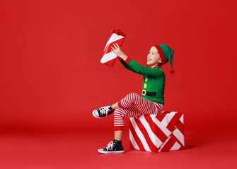 Sticker - cheerful funny child in Christmas elf costume with gifts on   red background.