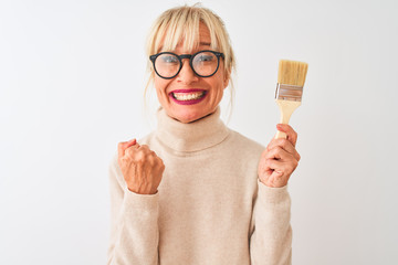 Sticker - Middle age painter woman holding paint brush standing over isolated white background screaming proud and celebrating victory and success very excited, cheering emotion