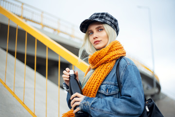 Portrait of young blonde teenager girl, wearing blue denim jacket, with orange scarf and plaid cap, holding a black steel thermo eco water bottle.