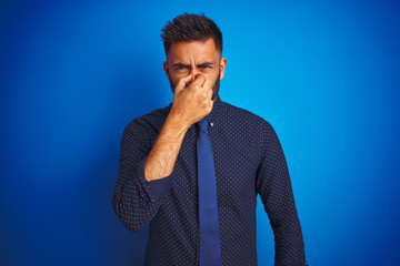 Canvas Print - Young indian businessman wearing elegant shirt and tie standing over isolated blue background smelling something stinky and disgusting, intolerable smell, holding breath with fingers on nose. 