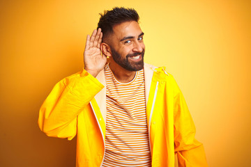 Poster - Young indian man wearing raincoat standing over isolated yellow background smiling with hand over ear listening an hearing to rumor or gossip. Deafness concept.