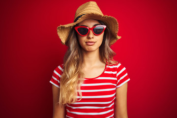 Sticker - Young beautiful woman wearing sunglasses and summer hat over red isolated background with serious expression on face. Simple and natural looking at the camera.