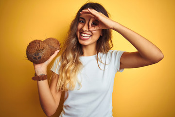 Wall Mural - Young beautiful woman holding coconut over yellow isolated background with happy face smiling doing ok sign with hand on eye looking through fingers