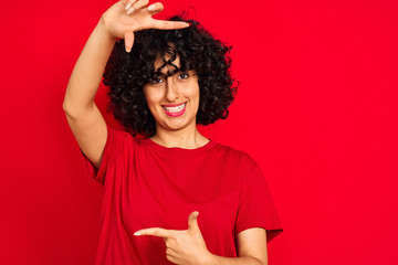 Sticker - Young arab woman with curly hair wearing casual t-shirt over isolated red background smiling making frame with hands and fingers with happy face. Creativity and photography concept.