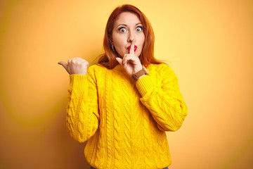 Wall Mural - Beautiful redhead woman wearing winter sweater standing over isolated yellow background asking to be quiet with finger on lips pointing with hand to the side. Silence and secret concept.