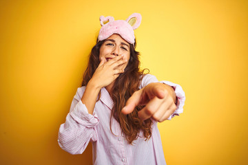 Wall Mural - Young woman wearing pajama and sleep mask standing over yellow isolated background laughing at you, pointing finger to the camera with hand over mouth, shame expression