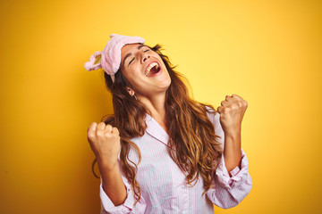 Wall Mural - Young woman wearing pajama and sleep mask standing over yellow isolated background celebrating surprised and amazed for success with arms raised and eyes closed. Winner concept.